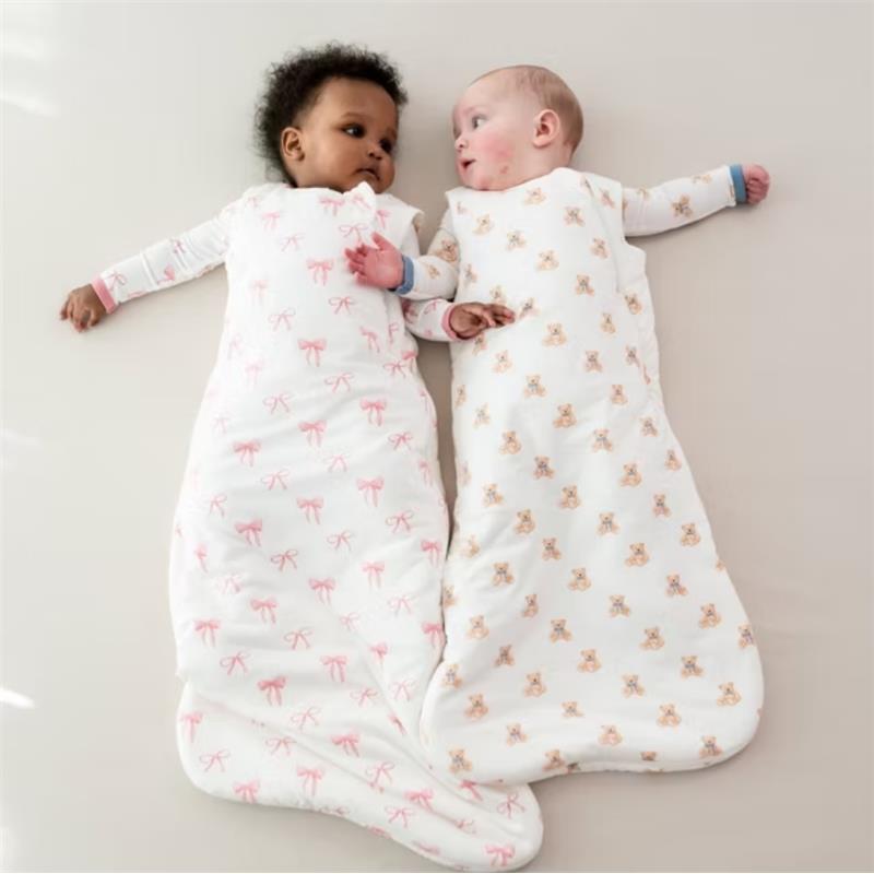 Two babies in Kyte Baby Sleep Bags: one with a pink bow design, the other with bear prints, lying side by side on a light surface.