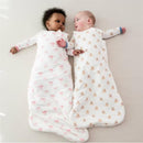 Two babies in Kyte Baby Sleep Bags: one with a pink bow design, the other with bear prints, lying side by side on a light surface.