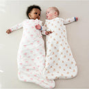 Two babies resting on a light surface, each in a Kyte Baby Sleep Bag; one features pink bows, the other has teddy bear prints.