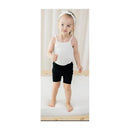 Toddler wearing a white spaghetti strap body and black shorts, standing on a bed with a neutral backdrop.