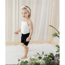 Toddler in a white spaghetti strap body and black shorts, smiling while standing on a light-colored surface with greenery around.