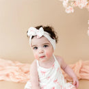 Toddler wearing a spaghetti strap ruffle romper with a pink bow print and matching headband, on a soft background.