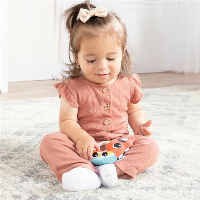 Toddler in a peach outfit sits on a rug, holding a colorful Lamaze Besties Rattle with animal faces.