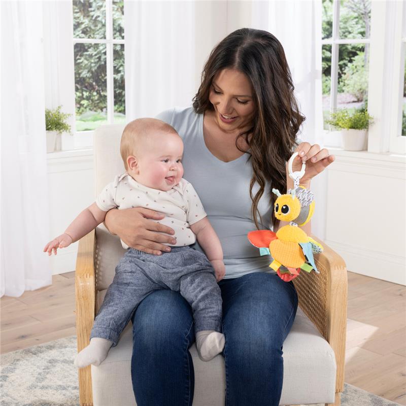 A woman holds a smiling baby while showcasing the Lamaze Buzzy The Bee Clip & Go toy, which is colorful and plush.