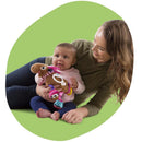 Baby holds a colorful Lamaze Clip On Car Seat and Stroller Toy, Muffin the Moose, while smiling with an adult in a green background.