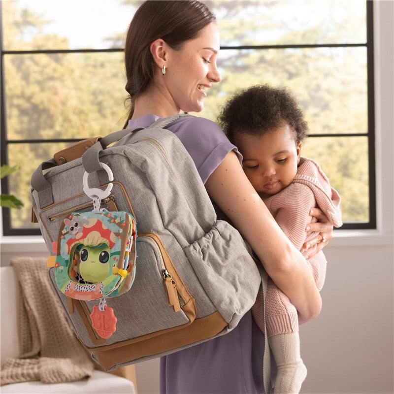 Gray and brown backpack with a colorful, plush baby toy attached; woman smiles while holding a child in a cozy setting.