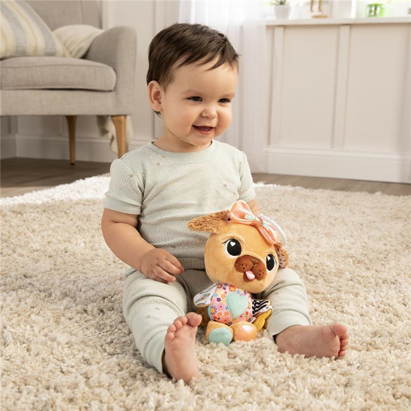Smiling toddler sitting on a soft rug, holding the Lamaze Lovey The Pug Clip & Go toy with colorful patterned accents.
