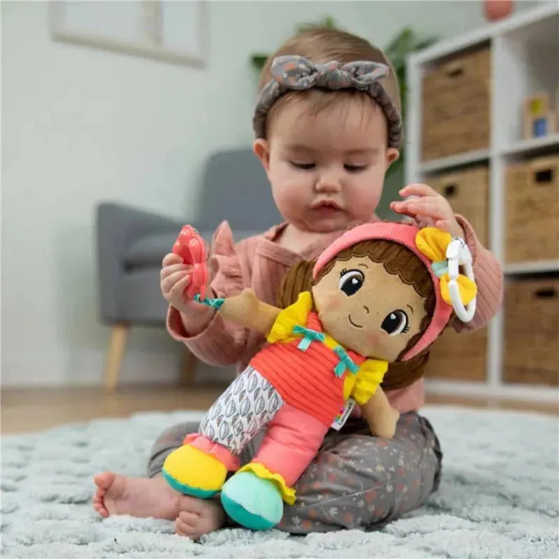 A baby sits on a rug, holding the Lamaze My Friend Jasmine sensory doll, wearing colorful clothes and a headband.