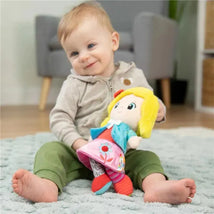 A smiling baby sits on a soft rug, holding the Lamaze My Friend Olivia doll, featuring bright colors and distinct patterns.