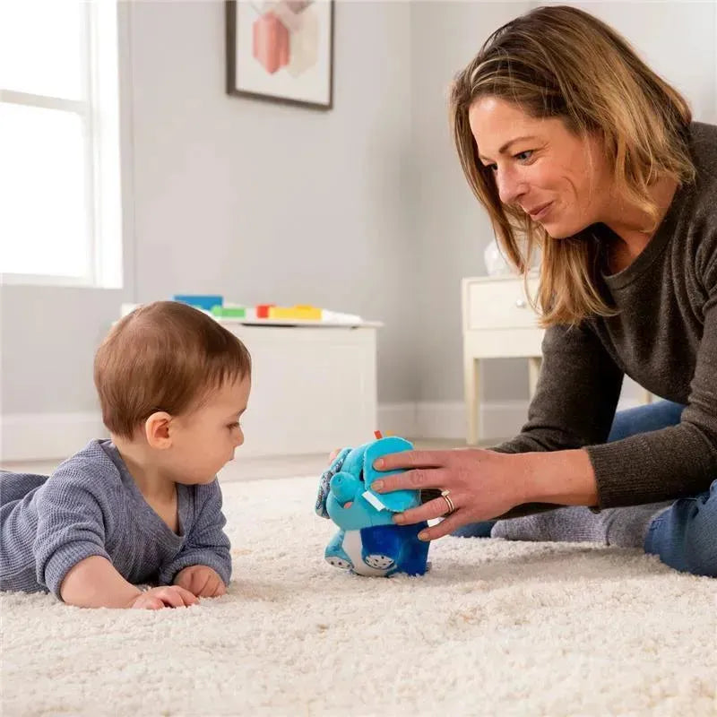 A toddler lies on a rug, focused on the Lamaze Puffaboo Elephant toy held by a smiling woman, enhancing sensory play.