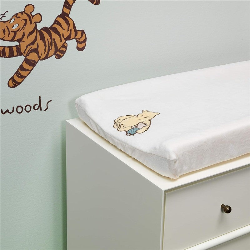 Changing pad cover featuring a whimsical illustration of Winnie the Pooh holding a blue blanket, displayed on a white changing table.