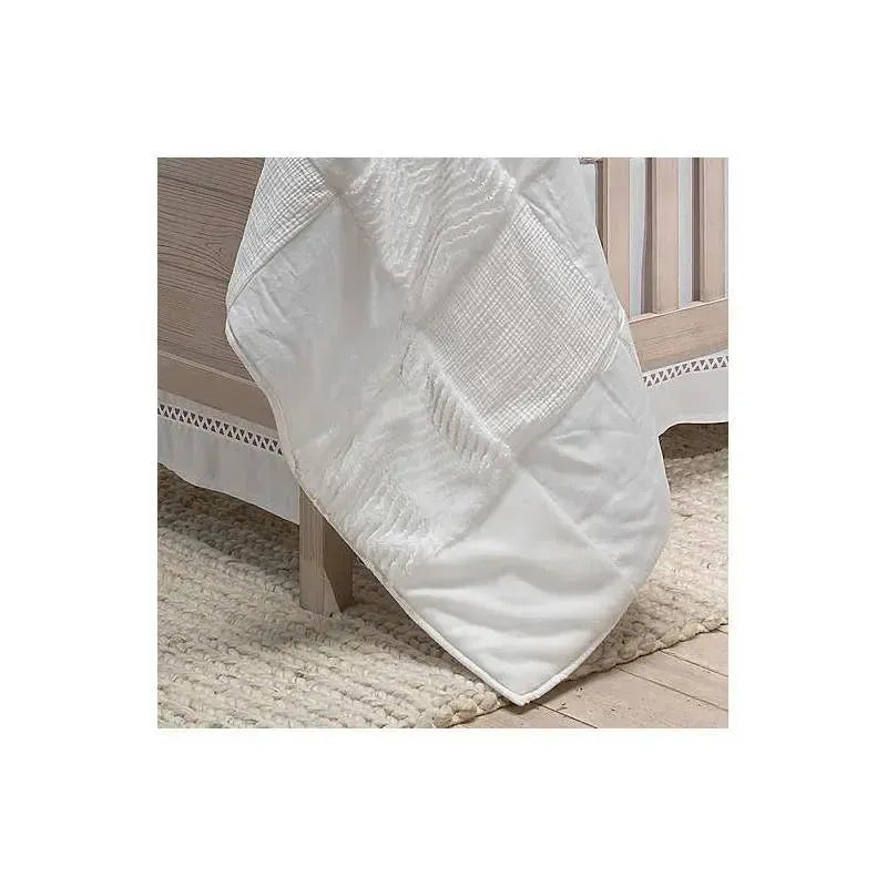 White patchwork quilt by Lambs & Ivy draped over a crib, featuring textured patterns and soft fabric details.