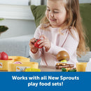 A young girl smiles while holding a red tomato toy, surrounded by colorful play food items and containers.