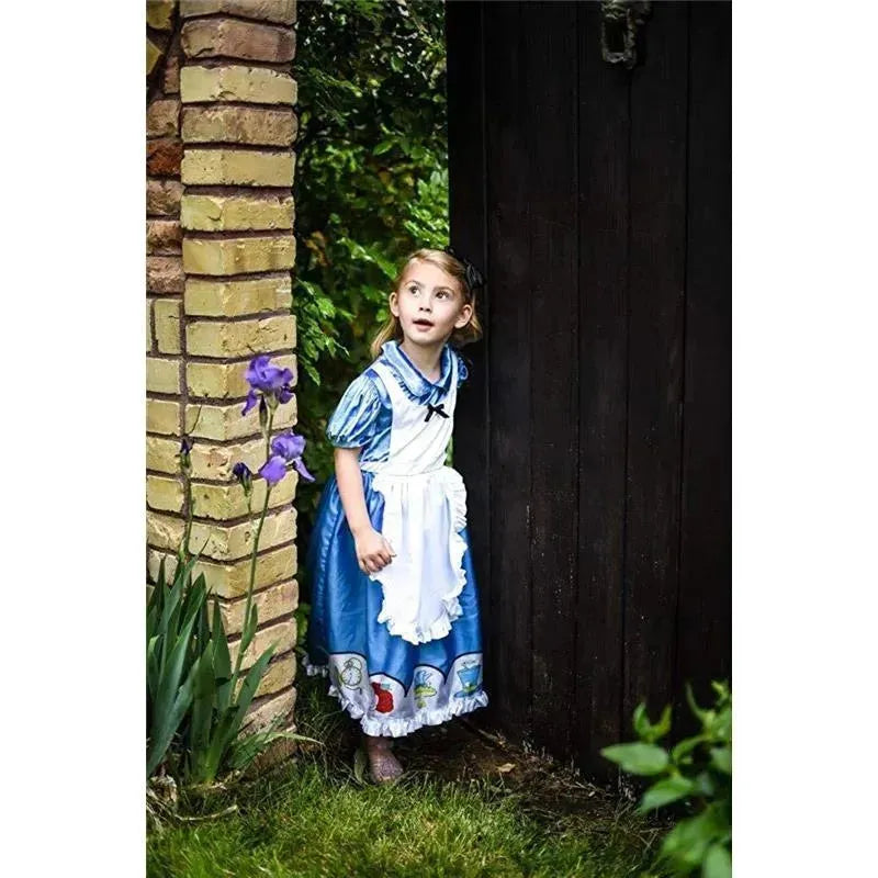 Young girl in a blue Alice in Wonderland dress peeks from behind a door, surrounded by greenery and purple flowers.