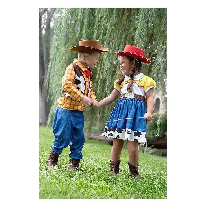 Children dressed in cowboy costumes hold hands and smile, surrounded by grass and trees. The boy wears a plaid shirt and cowboy hat, while the girl has a cow-print skirt and red hat.
