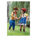 Children dressed in cowboy costumes hold hands and smile, surrounded by grass and trees. The boy wears a plaid shirt and cowboy hat, while the girl has a cow-print skirt and red hat.