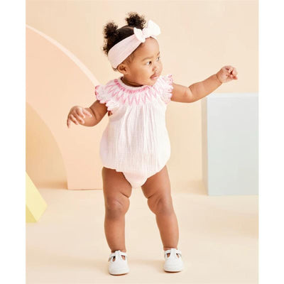 Baby girl wearing a pink smocked bubble outfit with ruffled sleeves, smiling and standing on a light-colored background.