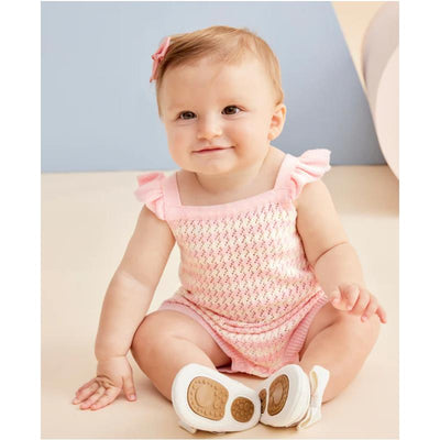 Baby girl in a pink striped bubble outfit with ruffled straps, sitting on a light-colored surface, smiling.