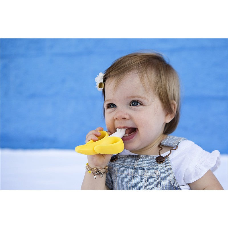 Infant happily uses a yellow Baby Banana Brush, designed for teething and brushing, against her gums.