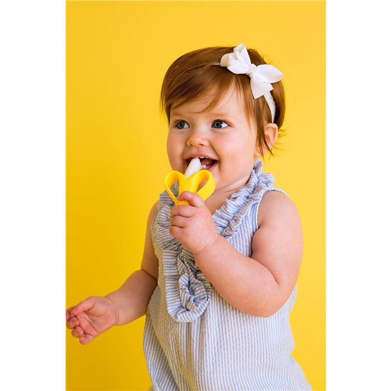 Infant smiling while holding a yellow Baby Banana Brush, designed for teething and oral care, against a bright yellow background.