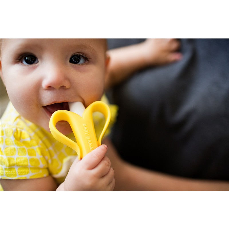 Infant holds a yellow banana-shaped brush, chewing on it, wearing a yellow patterned outfit.