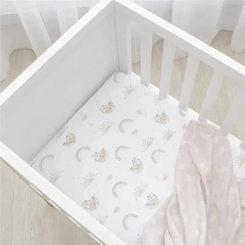 Cotton jersey crib fitted sheet featuring a rainbow sloth design, laid in a white crib with a soft pink blanket.
