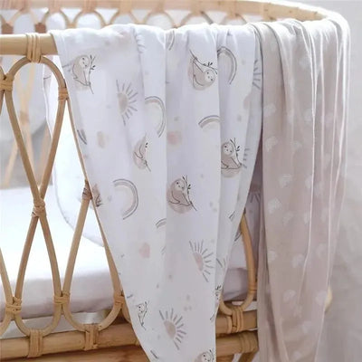 Two soft Jersey swaddles in a crib; one features sloths and rainbows, the other has a simple rainbow pattern.