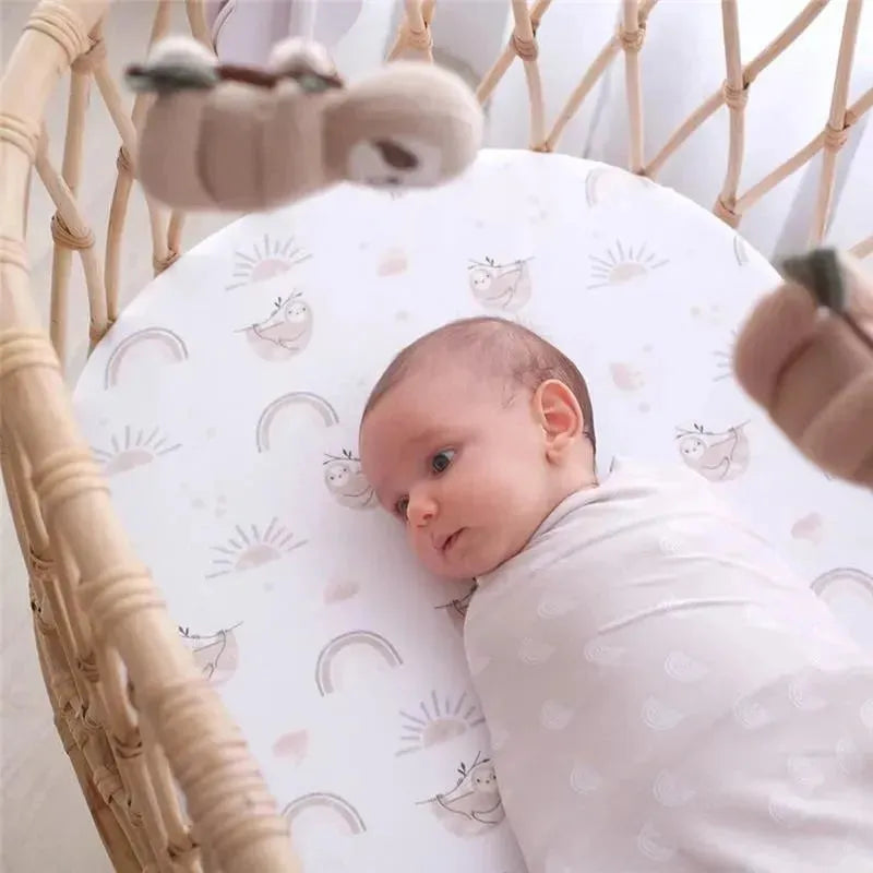 Baby wrapped in a soft swaddle, lying in a bassinet with a sloth and rainbow patterned mattress.
