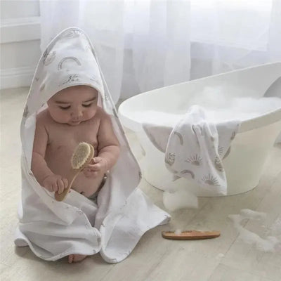 Baby wrapped in a white towel with rainbow patterns, playing with a wooden brush near a bubble-filled bathtub.