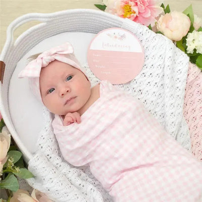 Newborn in a pink gingham wrap and headband, lying in a basket with flowers, beside an introduction card.