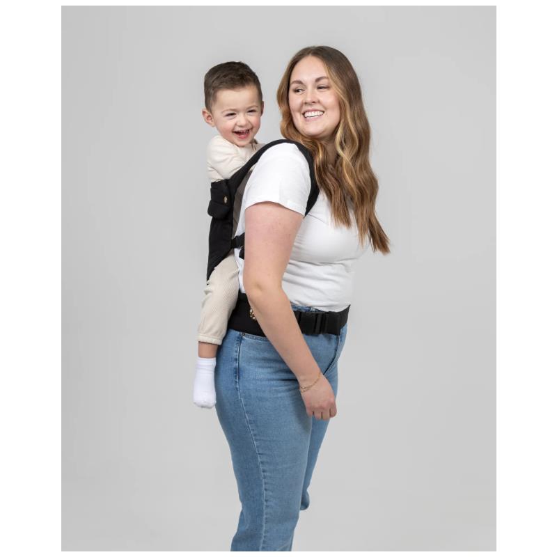 A woman wears the Mabe Monarch Carrier in Ebony, carrying a smiling child on her back, dressed in a white long-sleeve shirt and jeans.