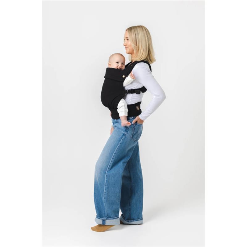 A woman in a white top carries a smiling baby in a black Mabe Monarch Carrier, wearing relaxed-fit jeans.