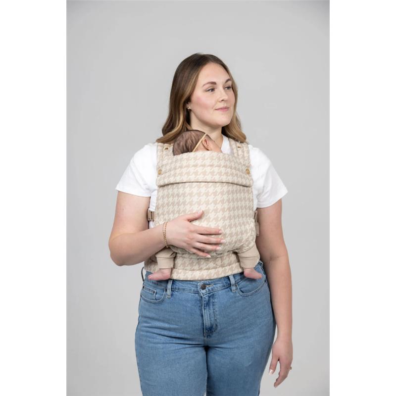 A woman wearing a houndstooth beige baby carrier, holding an infant securely against her chest.