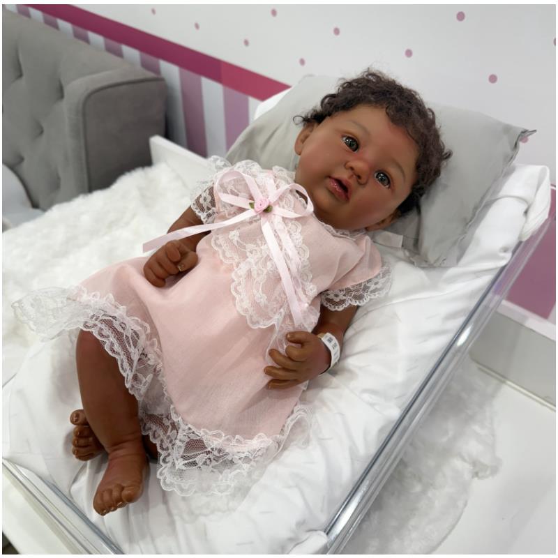 Reborn baby doll Shyann, an African American vinyl doll in a pink lace dress, lying on a pillow in a display crib.