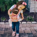 A child, wearing a pink hairbow, lovingly carries a doll in a matching pink maternity doll carrier, showcasing nurturing play.