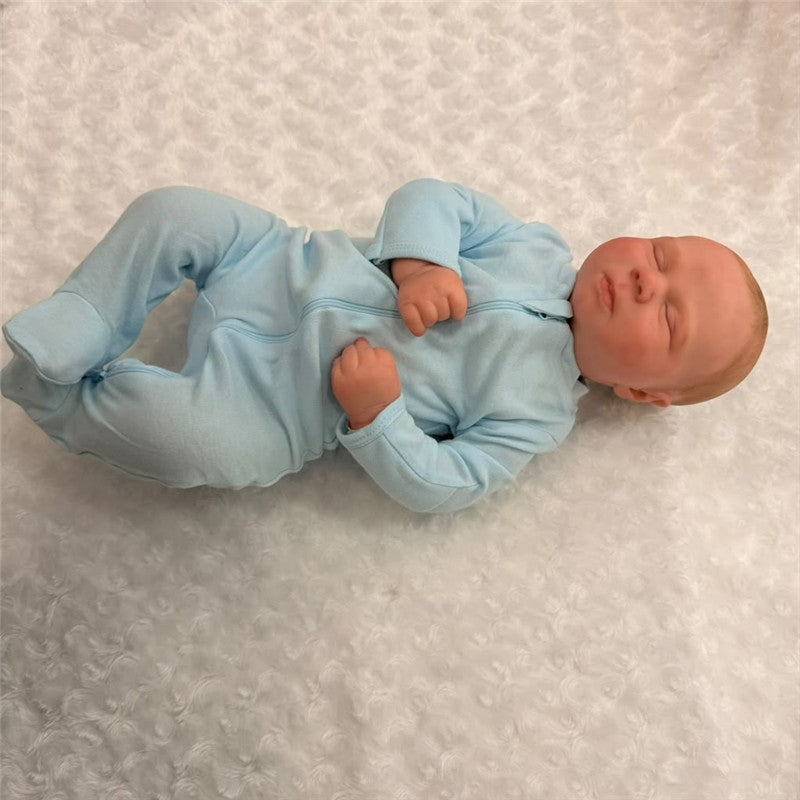 Macrobaby Reborn Doll named Charlie, dressed in a light blue onesie, lying peacefully on a soft white surface.