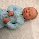 Macrobaby Reborn Doll Charlie dressed in a light blue onesie, resting peacefully on a textured white surface.