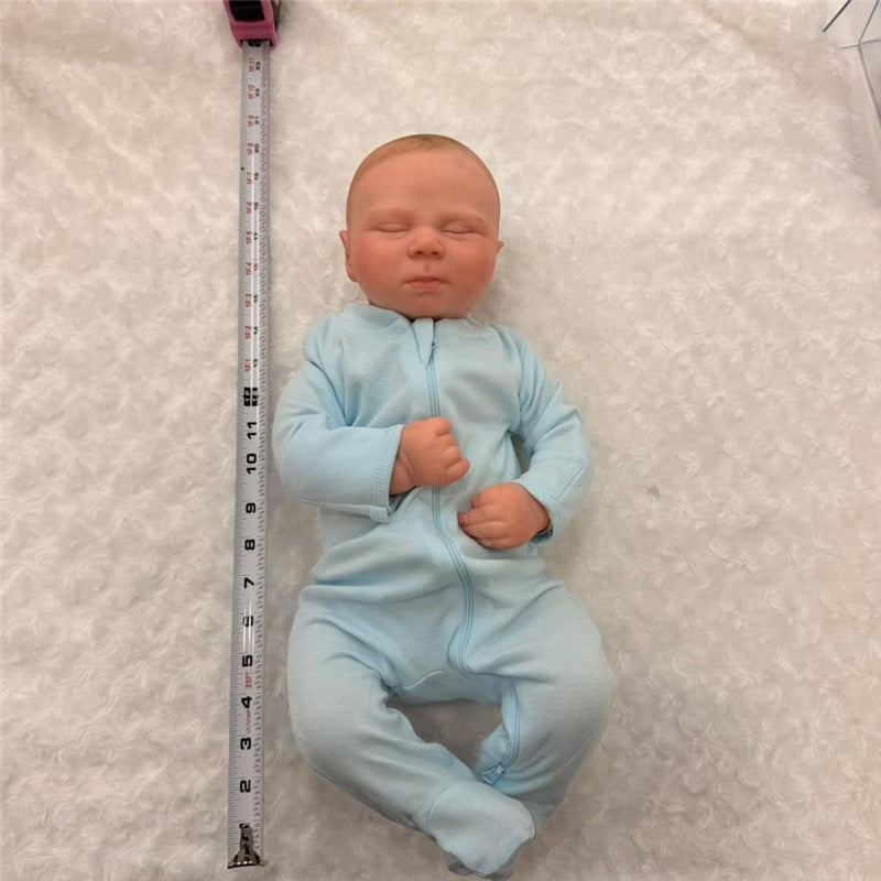 Reborn doll Charlie, made of white vinyl, wearing a light blue onesie, lying on a textured white surface with a measuring tape beside it.