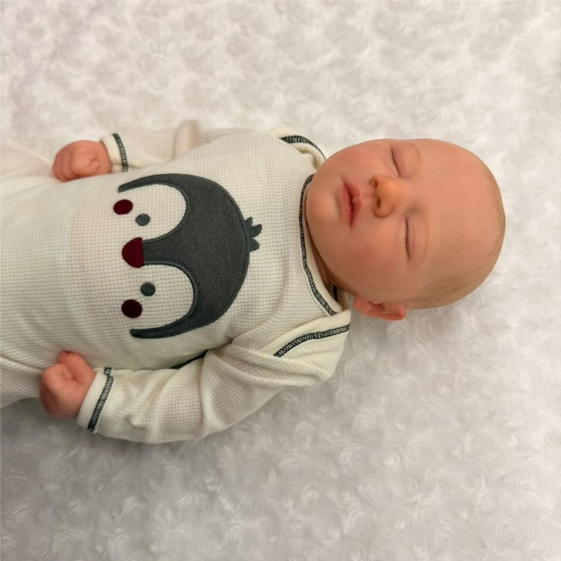 Reborn doll Jaxson Asleep 2 made of white vinyl, with painted hair and closed eyes, wearing a penguin-themed outfit.