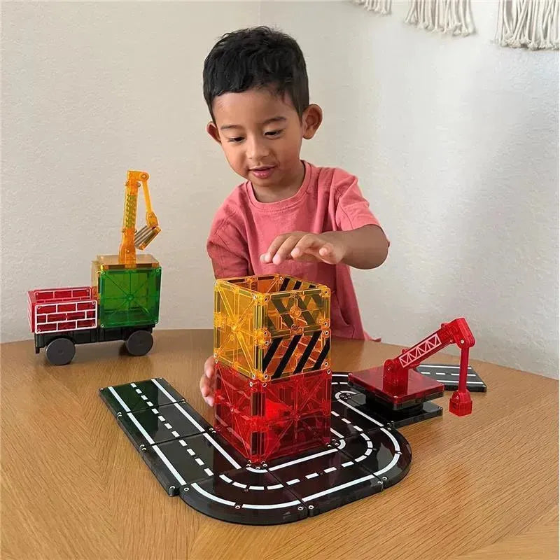 A child in a pink shirt builds a tall structure with colorful Magnatiles on a wooden table, featuring a toy truck and a crane.
