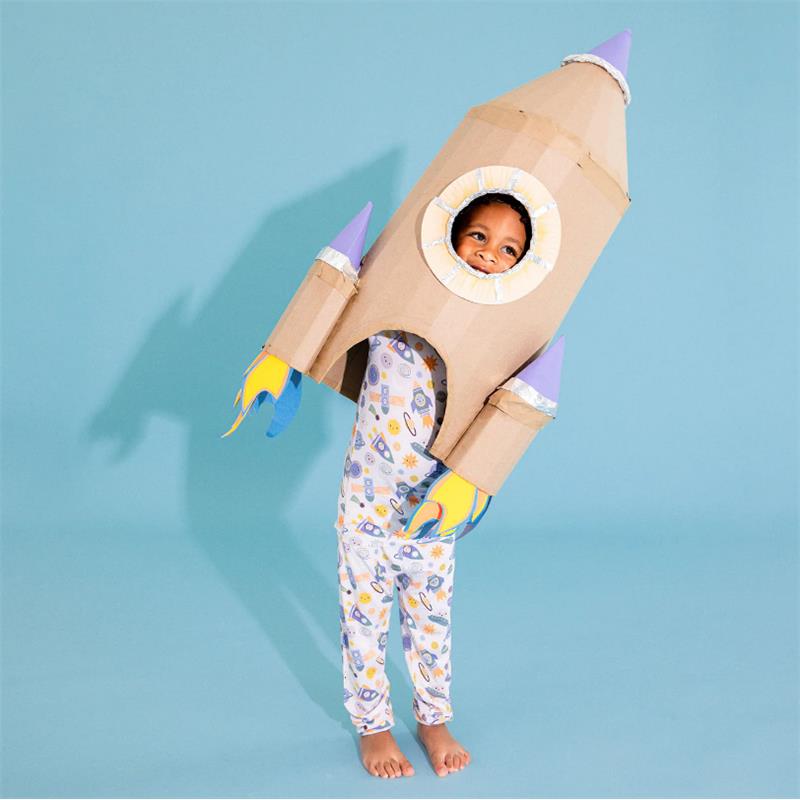 Child wearing colorful rocket-themed pajamas stands in a cardboard rocket costume, smiling against a light blue background.