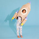 Child wearing colorful rocket-themed pajamas stands in a cardboard rocket costume, smiling against a light blue background.