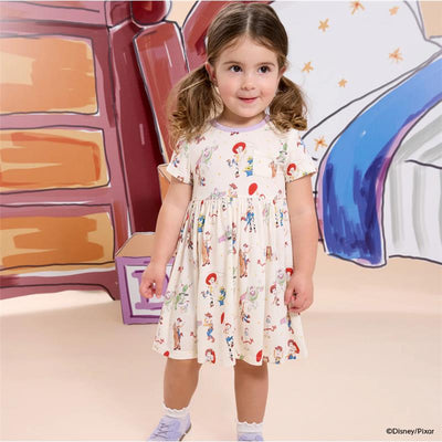 Toddler wearing a short sleeve dress featuring Toy Story characters, with a playful expression against a colorful background.