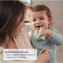 A toddler with curly hair holds a MAM baby toothbrush with a short handle, smiling while an adult looks on lovingly.