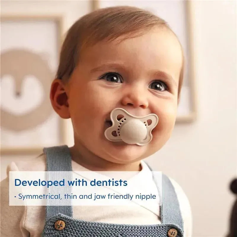 A smiling baby with a beige MAM Matte Deco pacifier in their mouth, wearing a light shirt and denim overalls.