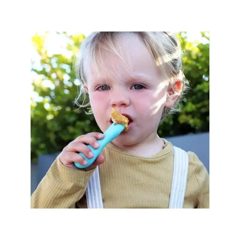 Child using a blue silicone palm grasp spoon holding food, wearing a beige shirt and white suspenders, outdoors.