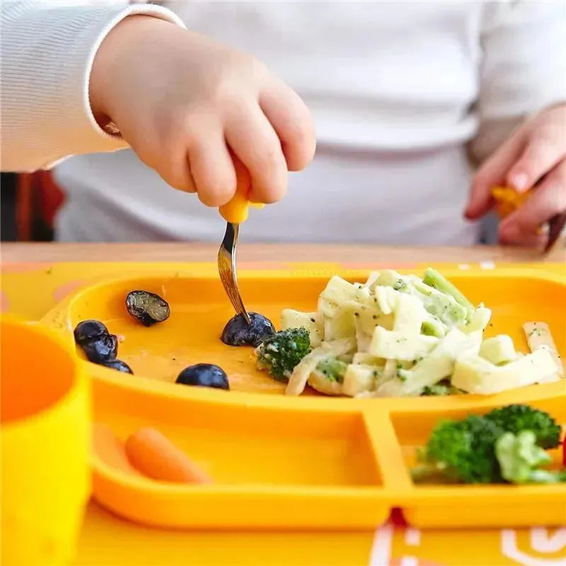 A toddler uses a yellow fork to pick up blueberries from a bright orange dining set, surrounded by pasta and broccoli.