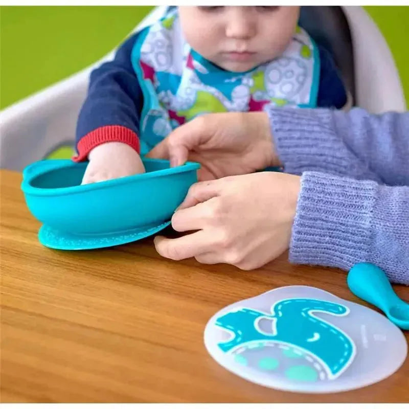 Toddler explores the Marcus & Marcus Lucas self-feeding set while an adult assists with a turquoise bowl and matching utensils on a wooden table.