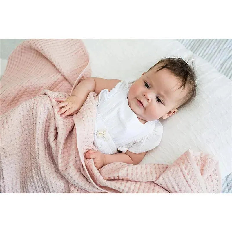 A baby wrapped in a pink Mary Meyer Lulujo Waffle Blanket with a textured pattern, lying on a white surface.