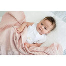 A baby wrapped in a pink Mary Meyer Lulujo Waffle Blanket with a textured pattern, lying on a white surface.